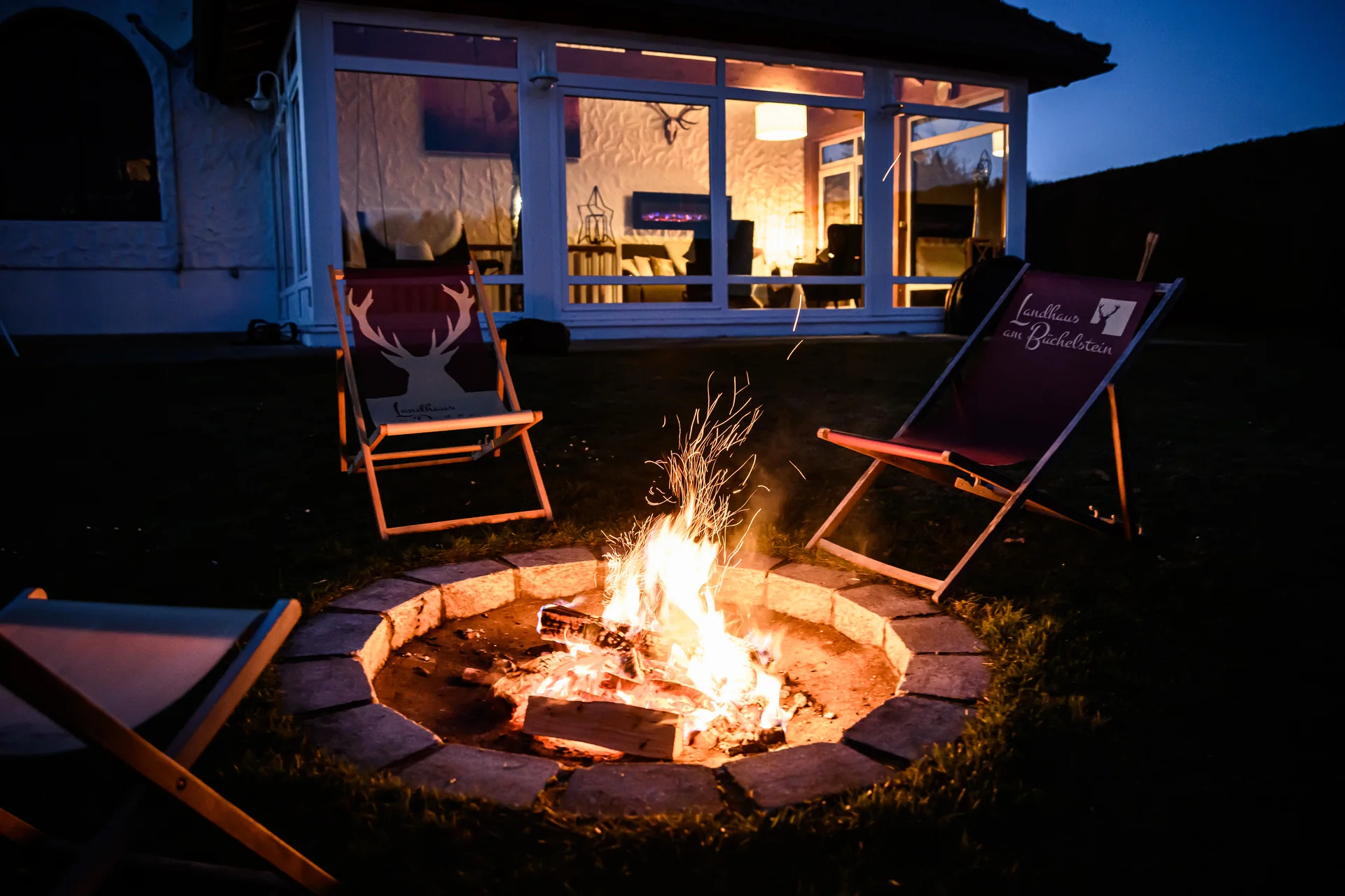 Abend im Garten des Landhauses mit Feuerstelle und dem warm beleuchteten Wintergarten dahinter